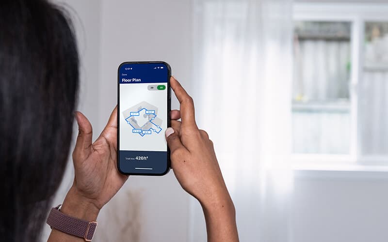 Hands holding a phone displaying a measured floor plan of the room around them.