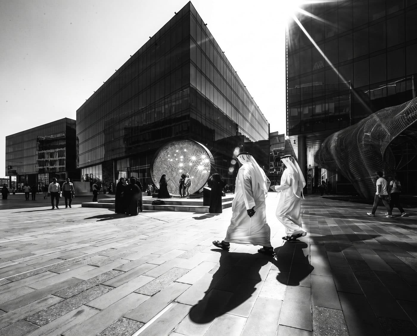 Two men in Emirati dress walking through a glass-fronted business district plaza, in black and white