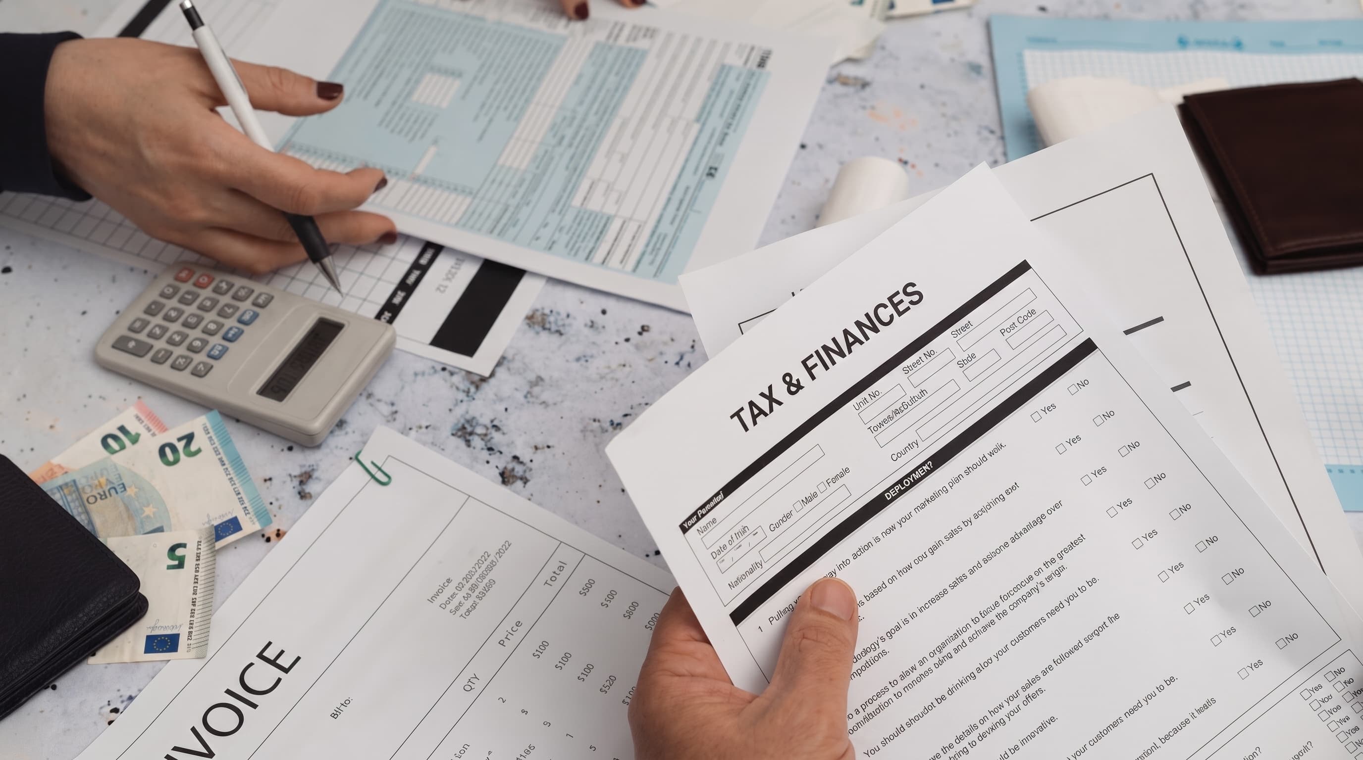 Hands reviewing tax and invoice paperwork beside a calculator and cash on a desk.